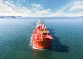 Large red oil tanker ship sailing on the ocean, blue sky with white clouds, mountains in the background.