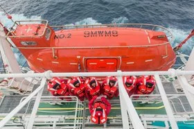 Maritime crew in red safety suits preparing near an emergency lifeboat on a ship.